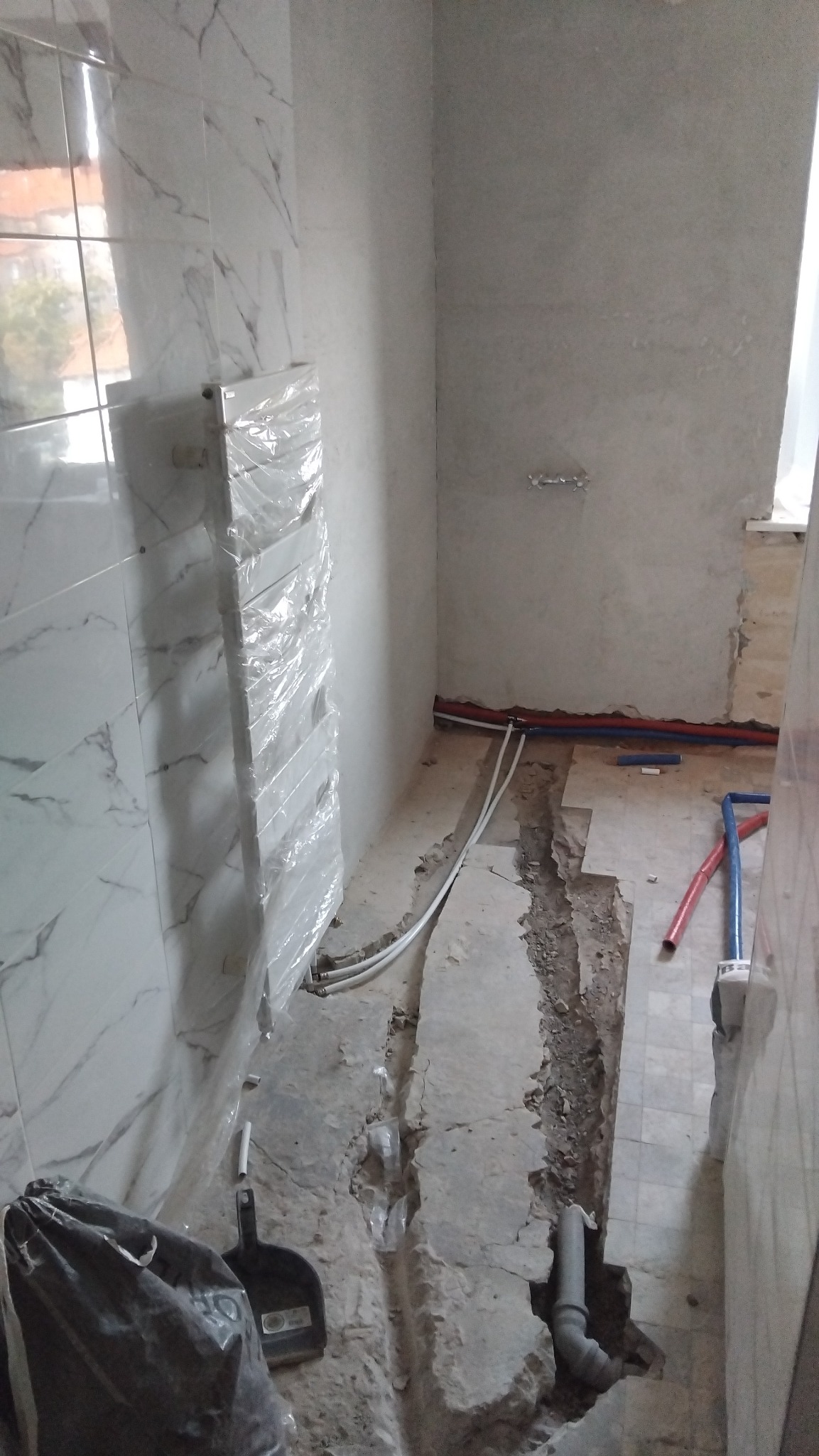 Remont łazienki: grzejnik owinięty folią, odkryte rury w bruzdach w podłodze, widoczne narzędzia. Etap prac hydraulicznych przed wykończeniem.