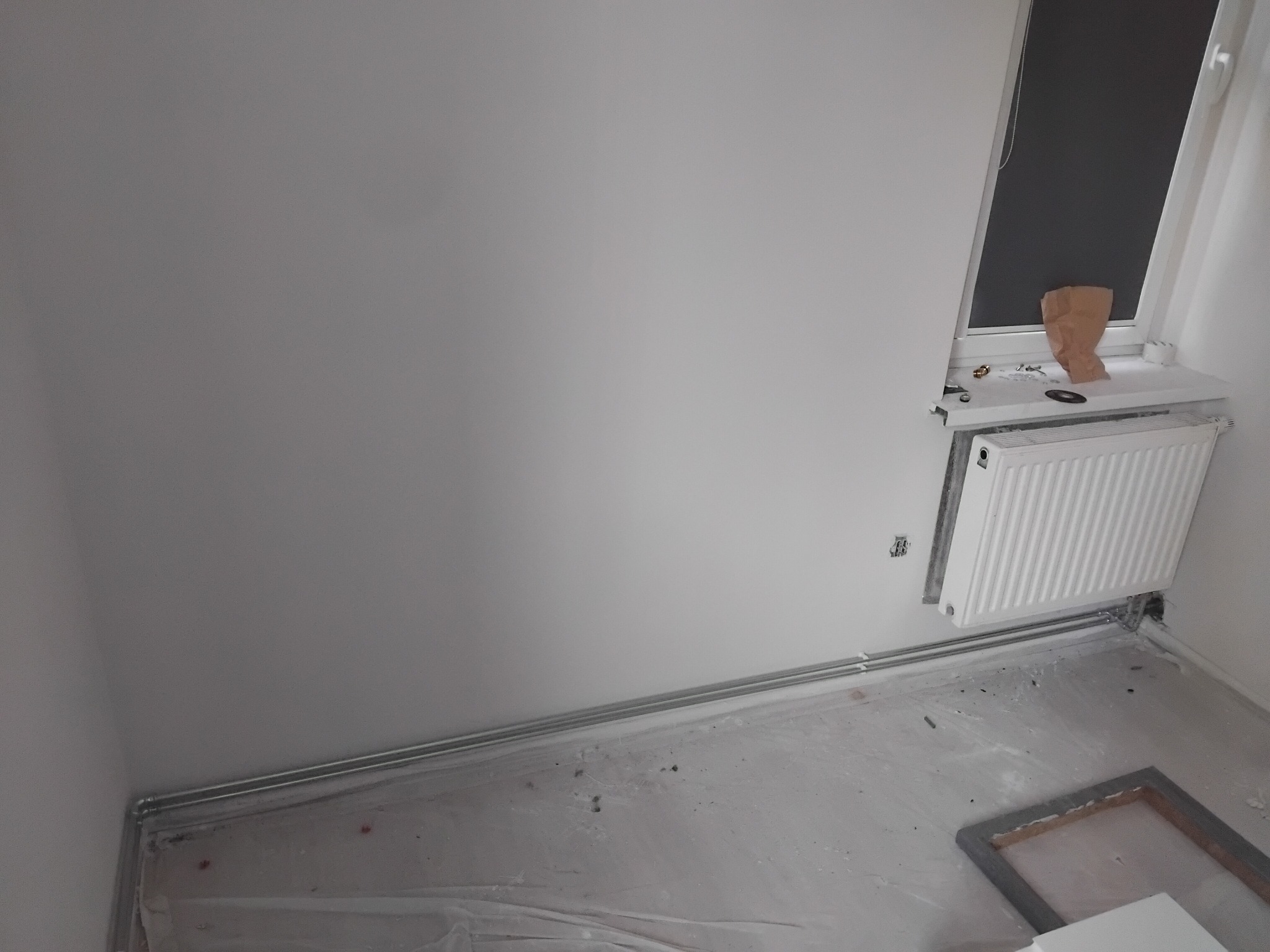 Biały grzejnik panelowy zamontowany pod oknem na tle białej ściany w trakcie remontu. Widoczne rury grzewcze i folia ochronna na podłodze.