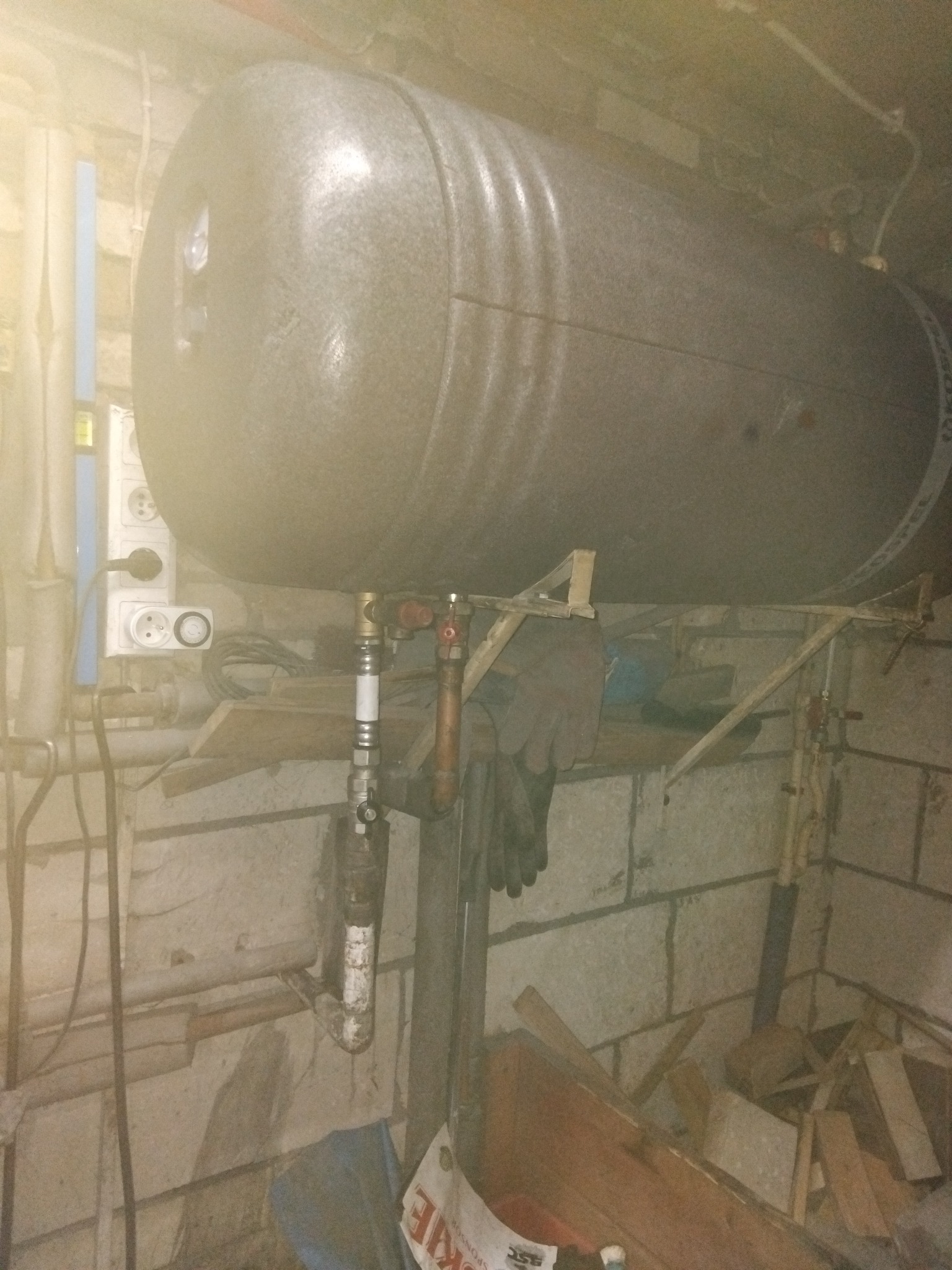 Szary, poziomy zbiornik na wodę z podłączonymi rurami i zaworami, zamontowany w piwnicy na prowizorycznej konstrukcji z drewna. Widoczne gniazdka elektryczne i fragment ściany z cegły.