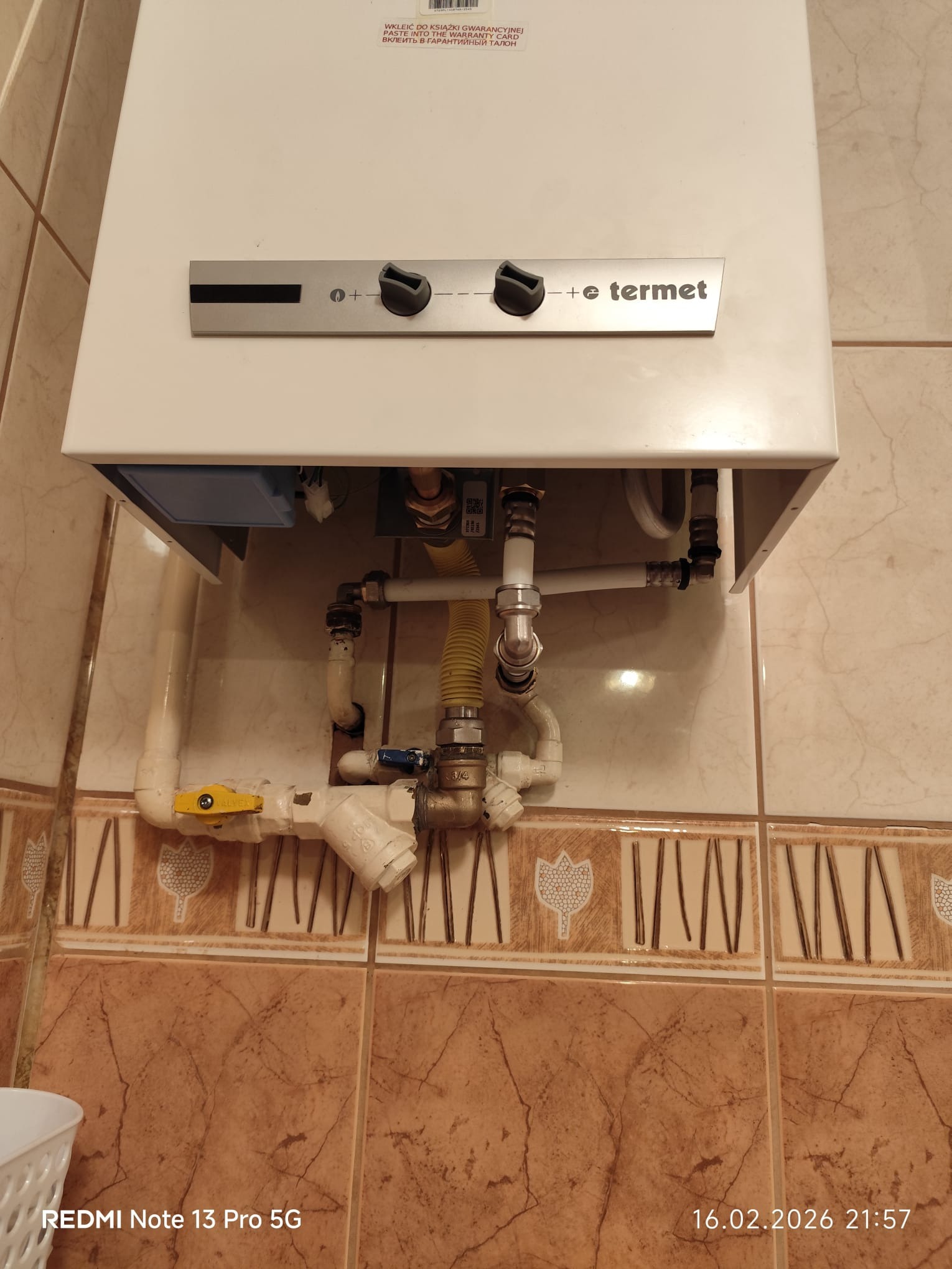 Biały piec gazowy Termet zamontowany na ścianie z brązowych płytek. Widoczne podłączenia hydrauliczne i gazowe. Żółty zawór odcinający gaz. Ujęcie z dołu.