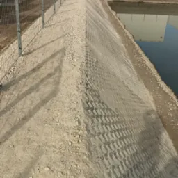 Skarpa ziemna umocniona ażurowymi płytami betonowymi, zabezpieczona siatką, z widocznym ogrodzeniem panelowym i odbiciem budynku w wodzie.