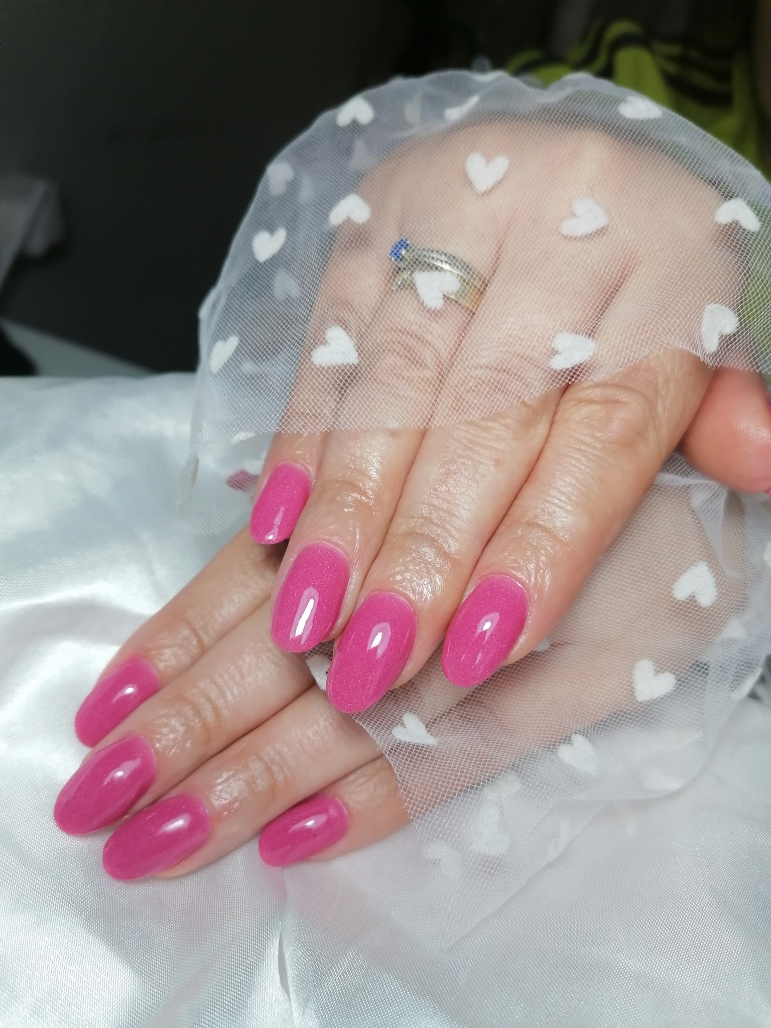 Dłonie z świeżo pomalowanymi, błyszczącymi paznokciami w odcieniu fuksji, ułożone na białej, delikatnej tkaninie w serduszka, pierścionek z niebieskim oczkiem na palcu wskazującym.