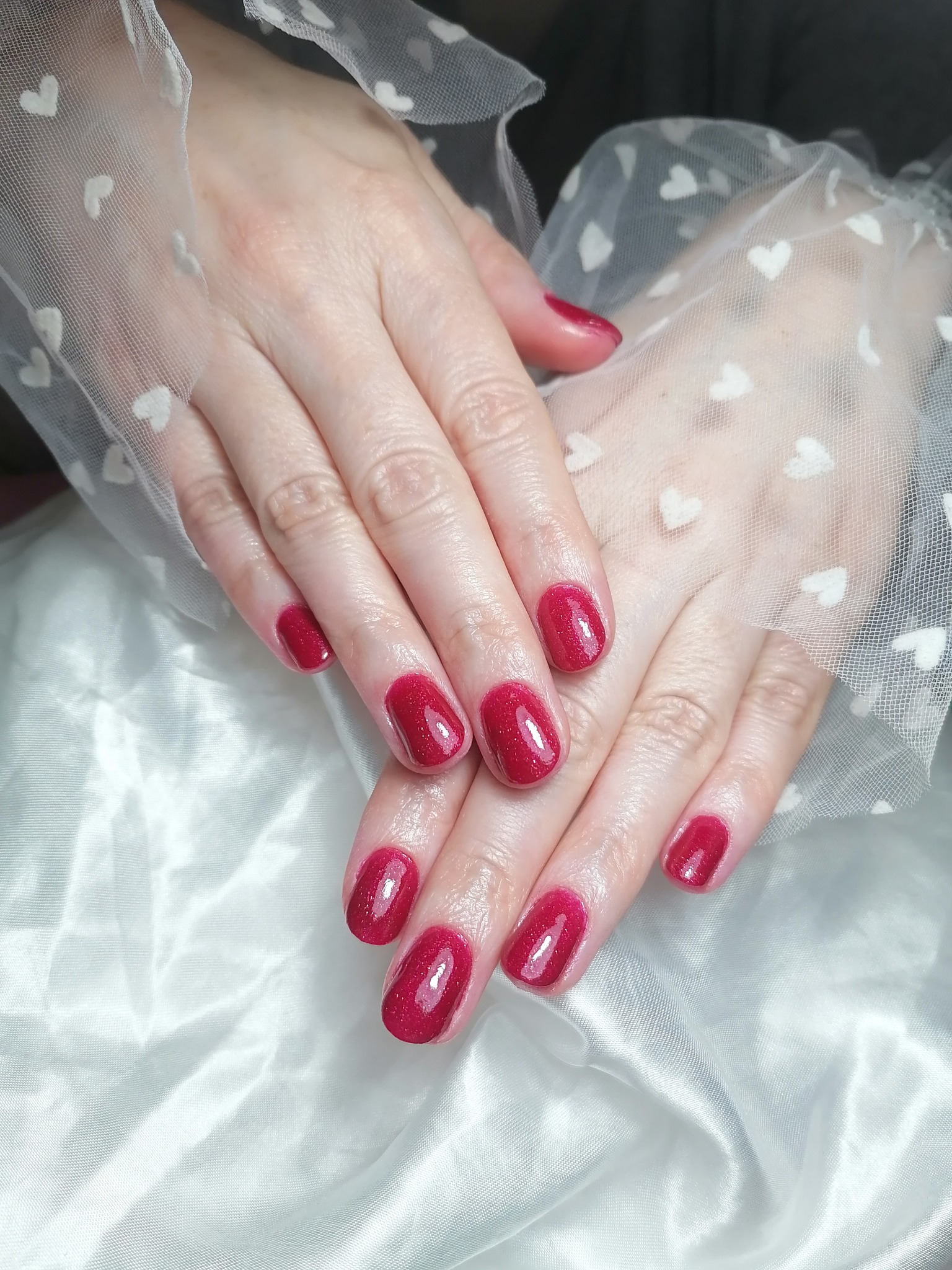 Dłonie z czerwonym, błyszczącym lakierem na krótkich paznokciach, ułożone na białej, satynowej tkaninie i przykryte tiulową rękawiczką w białe serduszka.