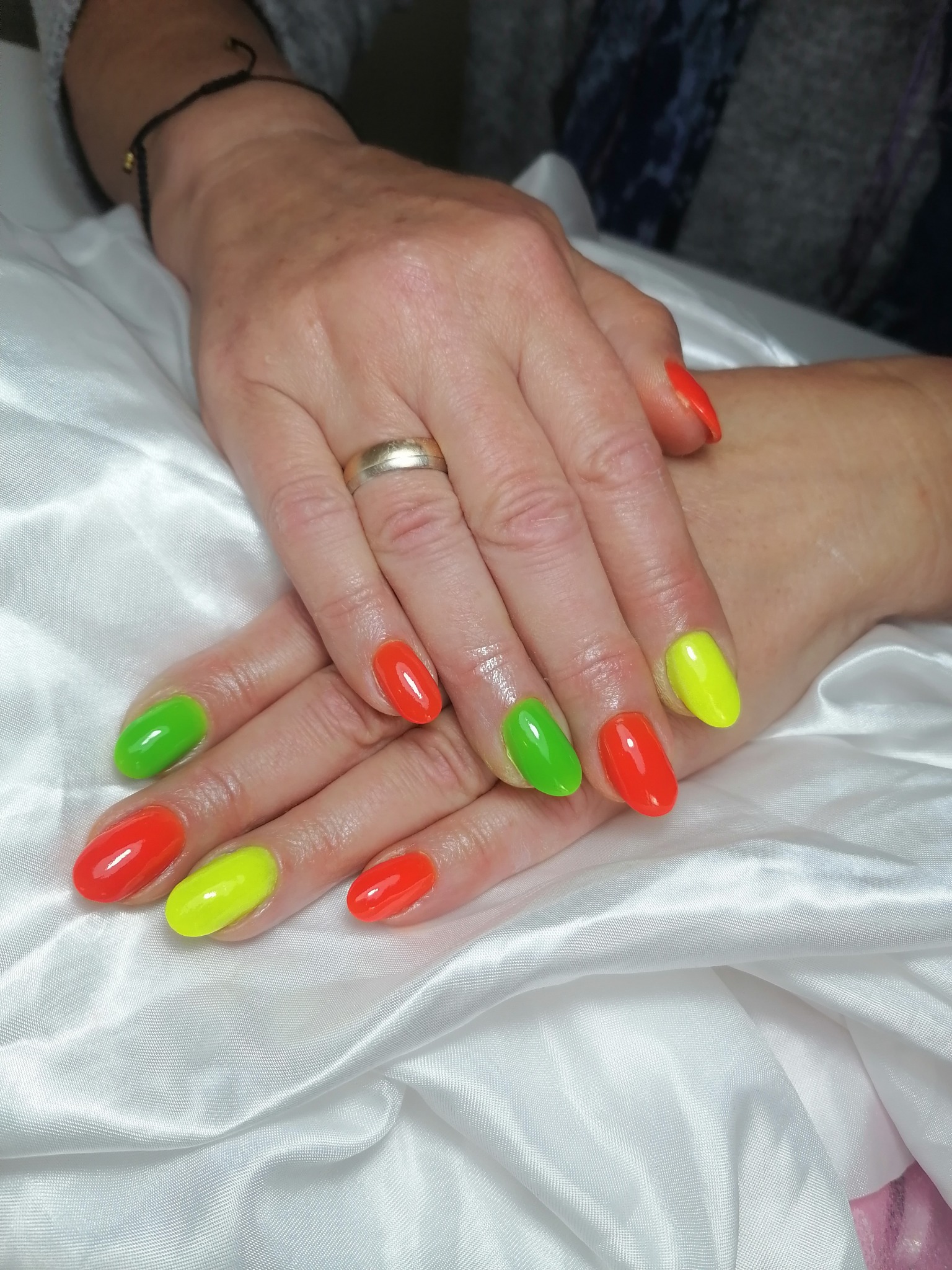 Dłonie z pomalowanymi paznokciami w kolorach czerwonym, zielonym i żółtym, ułożone na białej, satynowej tkaninie. Na palcu serdecznym widoczny złoty pierścionek.