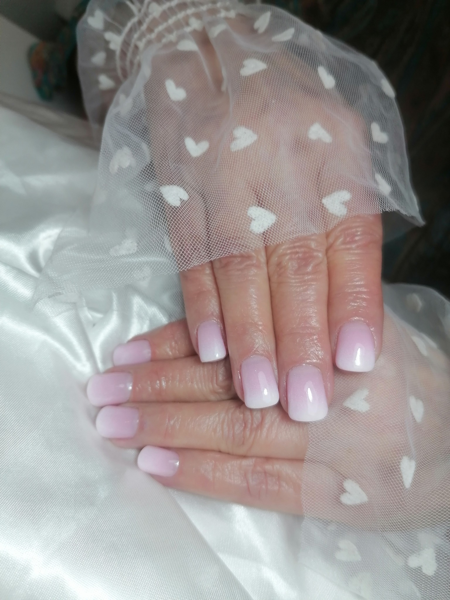 Dłonie z ombre manicure w odcieniach różu i bieli, ułożone na białej, satynowej tkaninie, przykryte tiulem w białe serduszka.