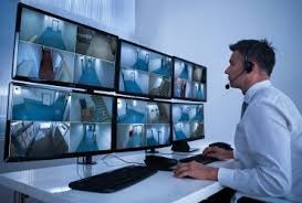 Mężczyzna w słuchawkach siedzi przed wieloma monitorami, na których wyświetlany jest obraz z kamer wewnątrz budynku. Widoczne korytarze i pomieszczenia biurowe.