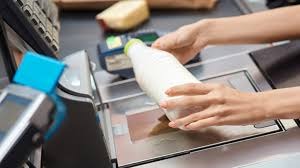 Kobieta skanuje butelkę mleka przy kasie samoobsługowej w supermarkecie, widoczny terminal płatniczy i skaner kodów kreskowych.