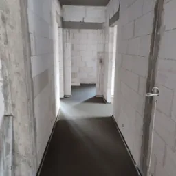 Świeżo wylana, gładka posadzka w surowym wnętrzu budynku z białych bloczków, widoczne ogrzewanie podłogowe i instalacje elektryczne na ścianach.