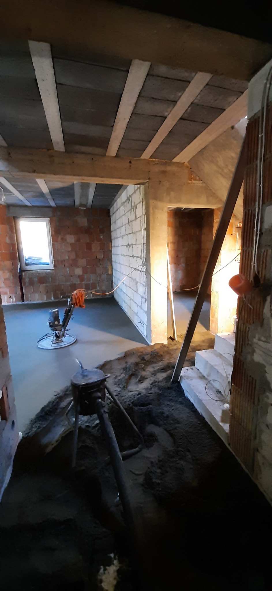 Wnętrze w trakcie wylewania posadzki jastrychowej z widoczną maszyną do zacierania oraz surowymi ścianami z cegły i betonu komórkowego.