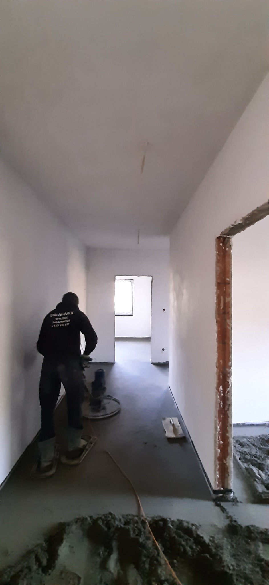 Aplikacja jastrychu cementowego w korytarzu budynku w stanie surowym, widoczny pracownik w ubraniu roboczym z logo firmy, zacierający powierzchnię mechaniczną zacieraczką, oraz resztki mokrej...