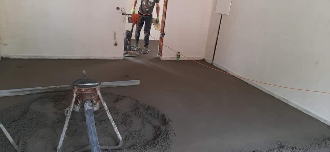 Wyrównywanie świeżo wylanego jastrychu maszyną zacieraczką w pomieszczeniu z widocznymi przewodami elektrycznymi i butelką wody przy ścianie.