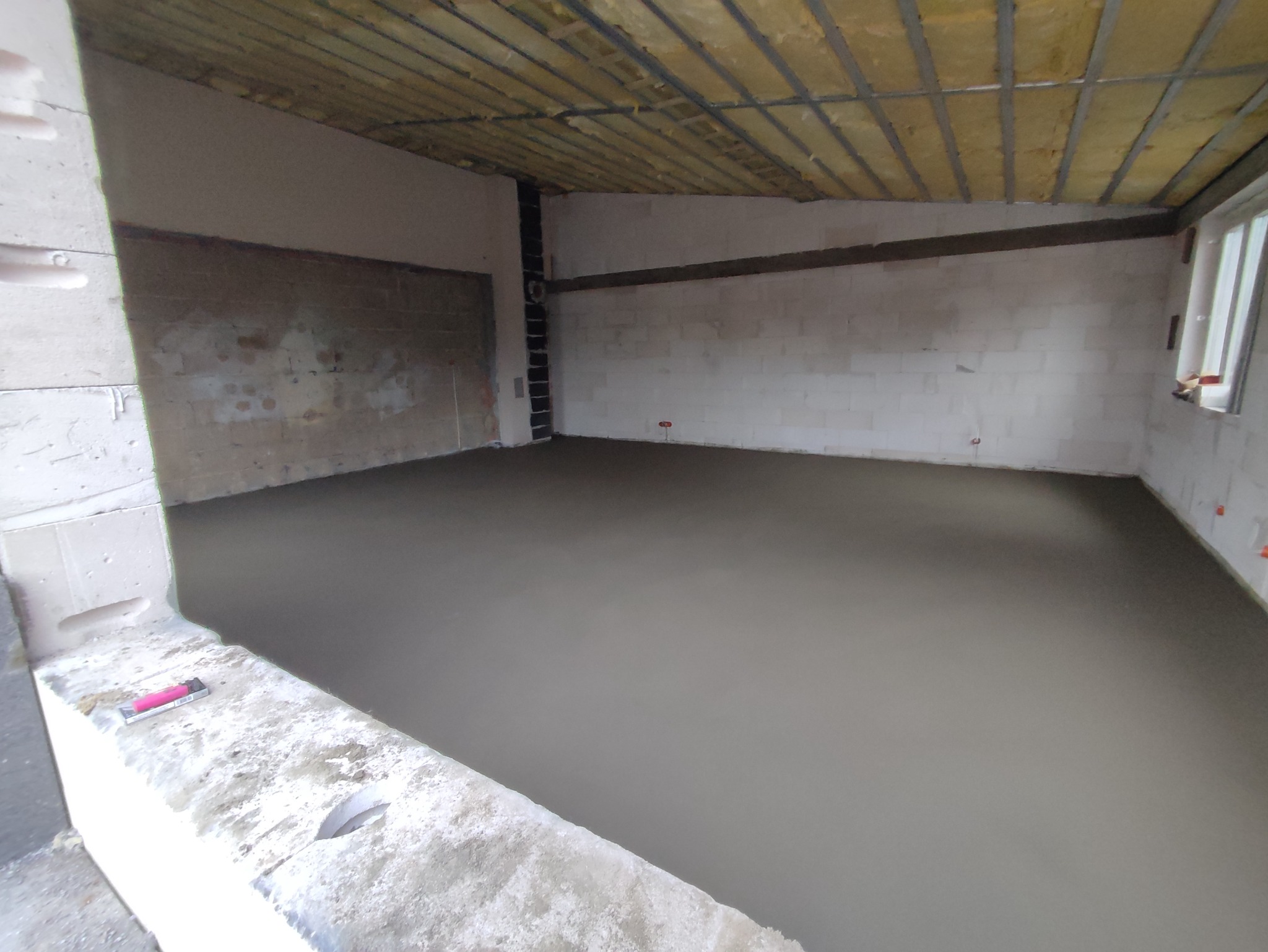 Wylany jastrych w surowym wnętrzu, widoczne ściany z betonu komórkowego i ocieplenie stropu, na pierwszym planie pozostawiony zapalniczka.