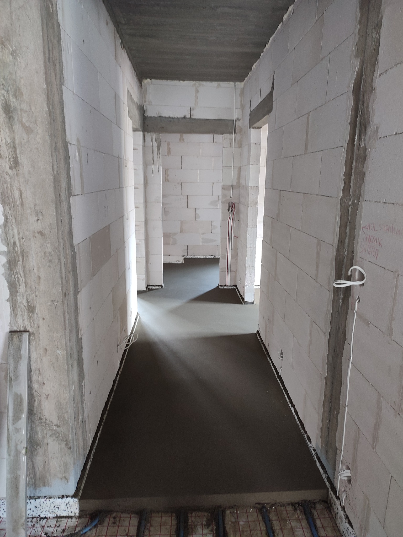 Świeżo wylana, gładka posadzka w surowym wnętrzu budynku z białych bloczków, widoczne ogrzewanie podłogowe i instalacje elektryczne na ścianach.