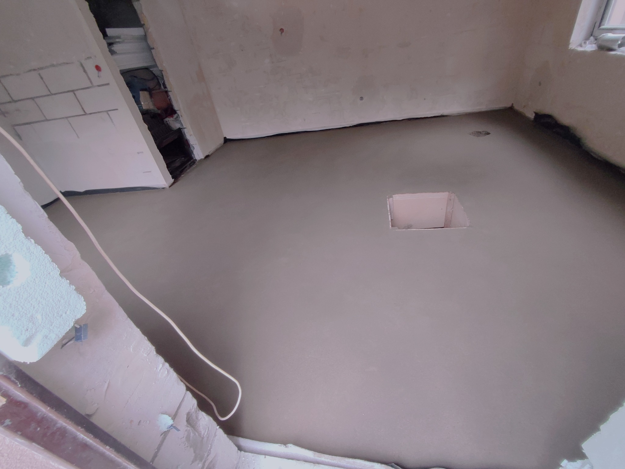 Świeżo wylana, gładka posadzka z jastrychu w trakcie wiązania, widoczny otwór rewizyjny w centralnej części pomieszczenia, ściany w stanie surowym.