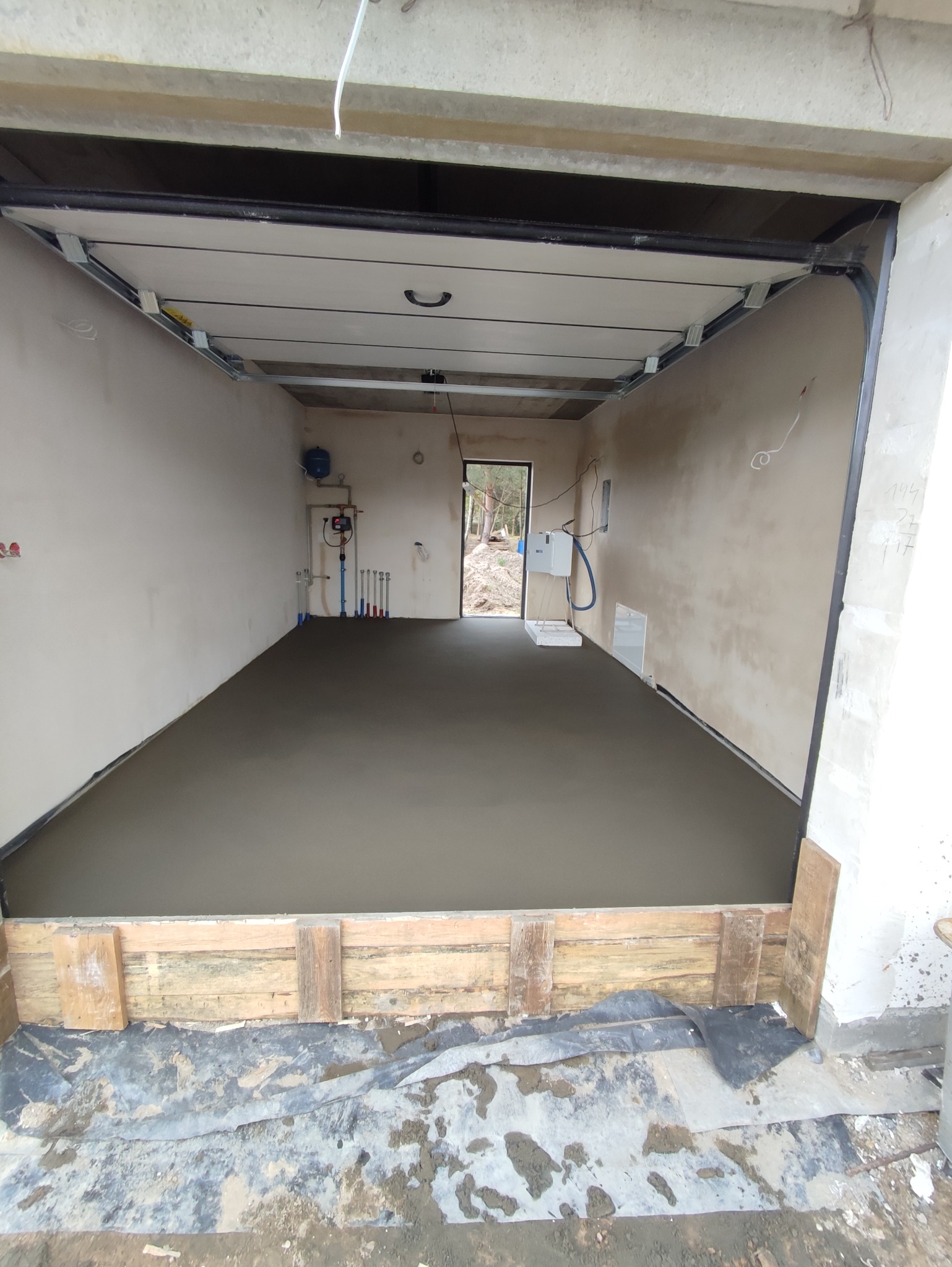 Świeżo wylana, gładka posadzka jastrychowa w garażu, z widocznymi deskami szalunkowymi przy krawędzi i instalacją grzewczą na ścianie w tle.