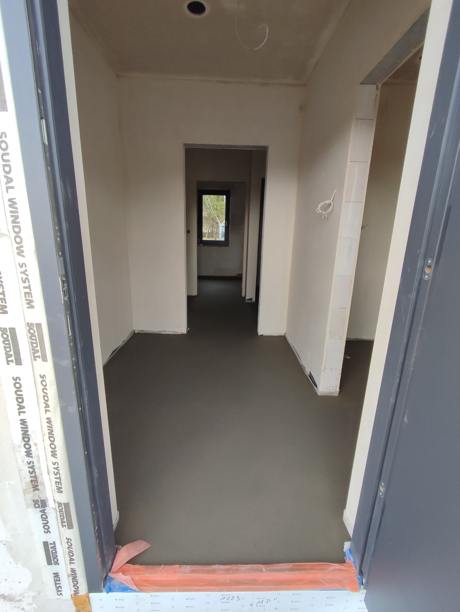 Świeżo wylana, szara posadzka jastrychowa w surowym wnętrzu z widocznymi przewodami elektrycznymi i oknem w oddali, zabezpieczona folią przy progu drzwi z widocznym napisem SOUDAL WINDOW SYSTEM.