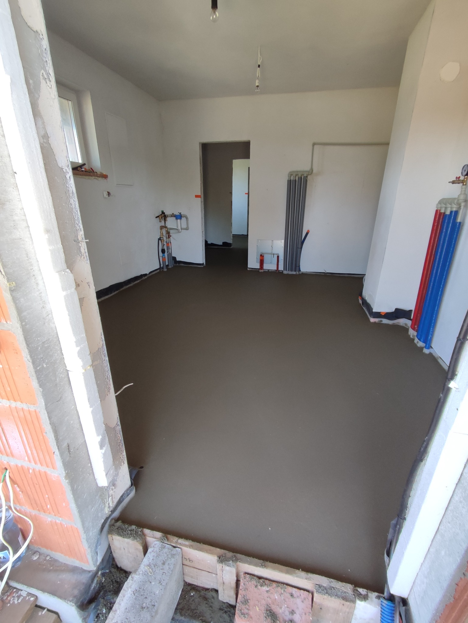 Wnętrze w trakcie prac wykończeniowych: wylana, świeża posadzka jastrychowa, widoczne instalacje grzewcze i wodne przy ścianach, surowe ściany w trakcie przygotowania do dalszych prac.