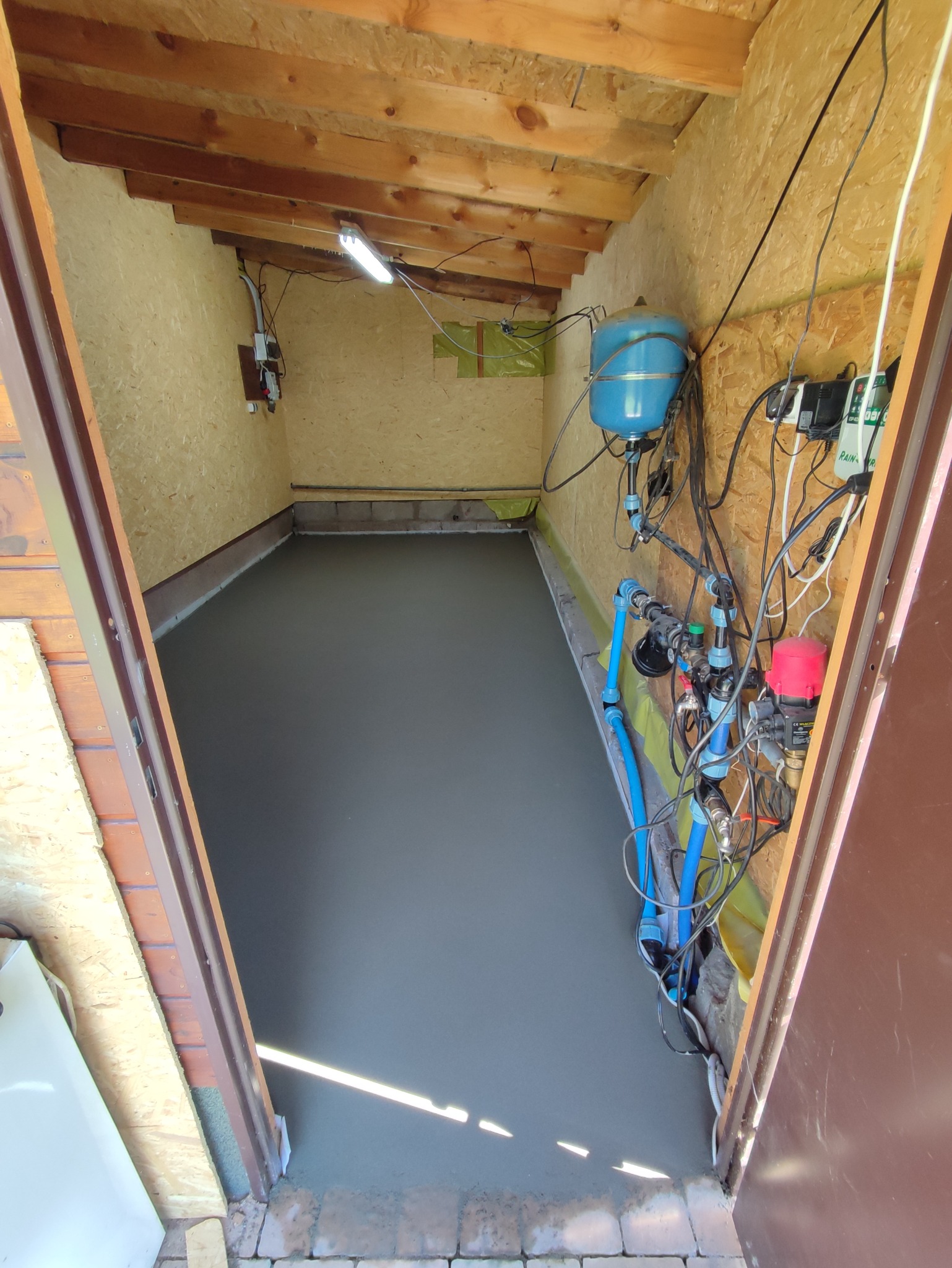 Świeżo wylana, gładka posadzka w pomieszczeniu gospodarczym z boazerią, widoczne instalacje wodne i elektryczne przy ścianie.