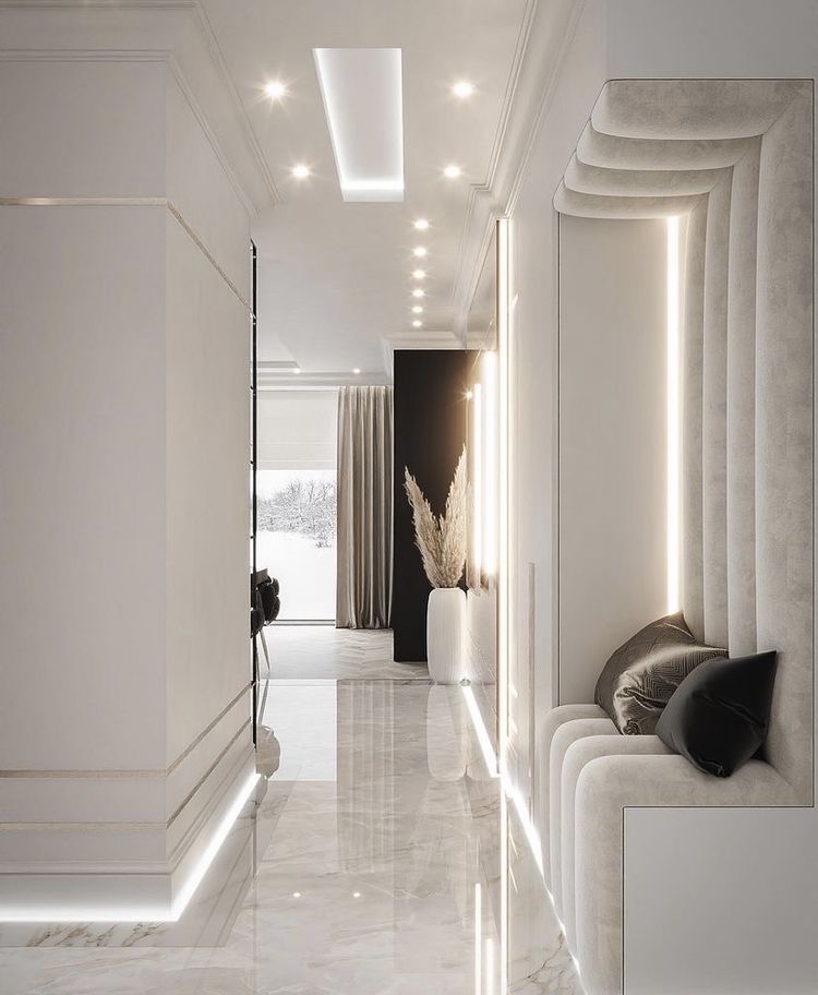 Nowoczesny korytarz z marmurową podłogą, oświetleniem LED wzdłuż ścian i sufitu, oraz wnęką z tapicerowanym siedziskiem i dekoracyjnymi poduszkami. W tle widoczny salon z zasłonami i widok...