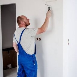 Preffer Home - Malarz w niebieskim kombinezonie maluje białą ścianę wałkiem, widoczne puszki instalacyjne w ścianie.