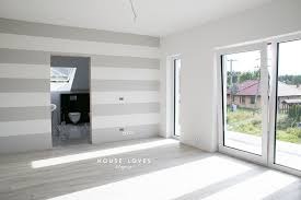 Jasne, przestronne wnętrze z poziomymi pasami w odcieniach szarości i bieli na ścianie. Widoczne duże okno balkonowe z widokiem na zieleń i zabudowania. W głębi pomieszczenia otwór drzwiowy...