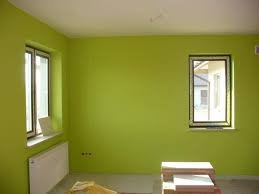 Pomalowane na jasnozielono wnętrze pokoju z dwoma oknami; widoczny grzejnik pod oknem po lewej stronie i niezabezpieczone gniazdka elektryczne na ścianie.