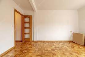 Pusty pokój z parkietem w jodełkę, świeżo pomalowane białe ściany i otwarte drzwi z przeszkleniami, kartonowe pudła w rogu.