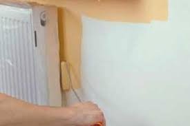 Malowanie ściany wałkiem przy grzejniku, zabezpieczone taśmą malarską, widoczna ręka malarza.