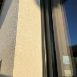 Fragment elewacji z jasnym tynkiem strukturalnym przy czarnej ramie okiennej, oświetlony popołudniowym słońcem z widocznym cieniem na rogu budynku.