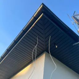 Nowoczesna elewacja domu z ciemnoszarą podbitką dachową i wystającymi przewodami do oświetlenia zewnętrznego, sfotografowana pod kątem w słoneczny dzień.