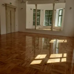 Parkiet ułożony w jodełkę francuską, świeżo po lakierowaniu, z widocznymi refleksami słonecznymi w przestronnym, jasnym wnętrzu z kolumnami i zielonymi ramami okiennymi.