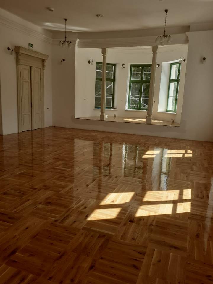 Parkiet ułożony w jodełkę francuską, świeżo po lakierowaniu, z widocznymi refleksami słonecznymi w przestronnym, jasnym wnętrzu z kolumnami i zielonymi ramami okiennymi.