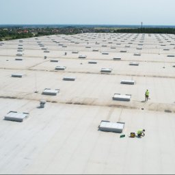 DACH-CONTROLL SPÓŁKA Z OGRANICZONĄ ODPOWIEDZIALNOŚCIĄ - Rozległy, płaski dach przemysłowy z licznymi świetlikami; dwóch pracowników w odblaskowych kamizelkach wykonuje prace naprawcze na jasnej powierzchni dachu, widoczne narzędzia i agregat prądotwórczy.