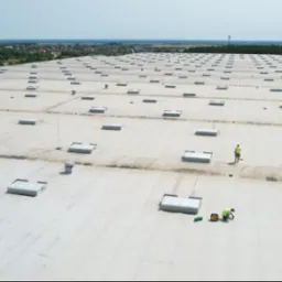 Rozległy, płaski dach przemysłowy z licznymi świetlikami; dwóch pracowników w odblaskowych kamizelkach wykonuje prace naprawcze na jasnej powierzchni dachu, widoczne narzędzia i agregat prądotwórczy.