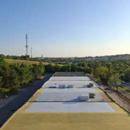 Widok z góry na rozległy dach budynku w trakcie prac izolacyjnych, z widoczną warstwą żółtej wełny mineralnej i białej membrany, na tle zielonego krajobrazu miejskiego i błękitnego nieba.