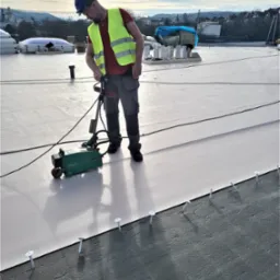 Dekarz w kasku i kamizelce odblaskowej zgrzewa membranę dachową na płaskim dachu za pomocą zgrzewarki VARIMAT V2, widoczne mocowania mechaniczne membrany.