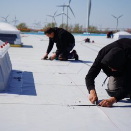 DACH-CONTROLL SPÓŁKA Z OGRANICZONĄ ODPOWIEDZIALNOŚCIĄ - Pracownicy na dachu z białej membrany, przycinają materiał nożem, w tle turbiny wiatrowe.