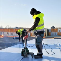DACH-CONTROLL SPÓŁKA Z OGRANICZONĄ ODPOWIEDZIALNOŚCIĄ - Dekarze zgrzewają membranę dachową na płaskim dachu za pomocą zgrzewarki automatycznej, w tle izolacja termiczna BauderPIR.
