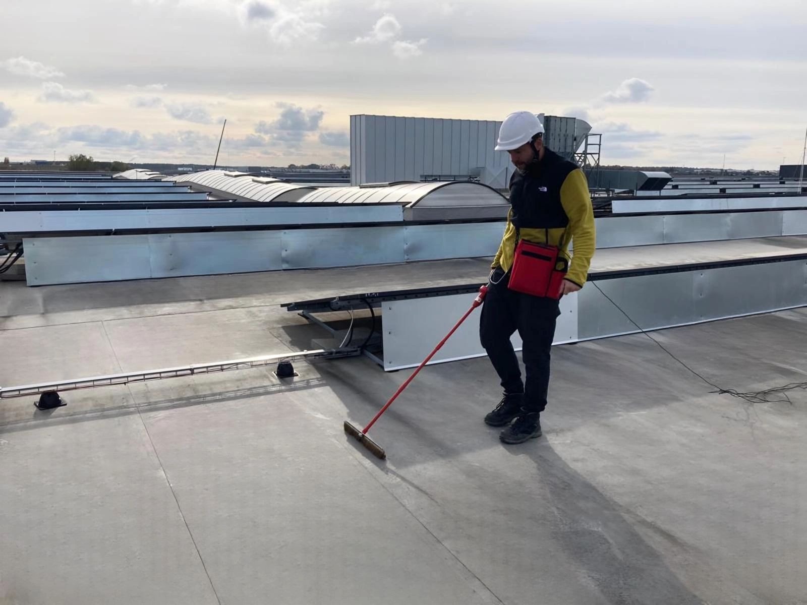 Pracownik w kasku i roboczej kurtce zamiata płaski dach przemysłowy z widocznymi elementami konstrukcyjnymi i świetlikami.