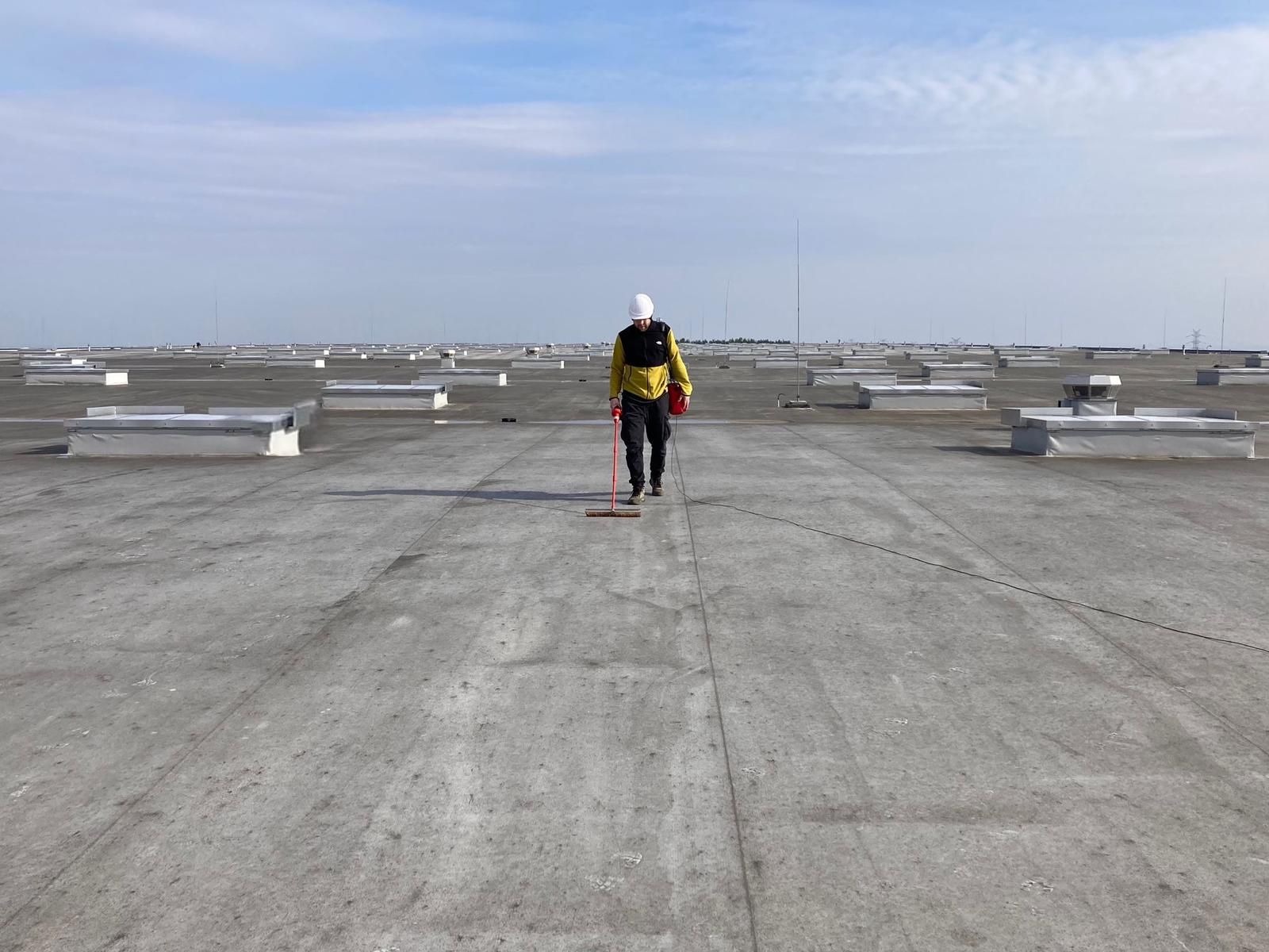 Pracownik w kasku i kurtce roboczej z narzędziem na rozległym, płaskim dachu przemysłowym z licznymi świetlikami.