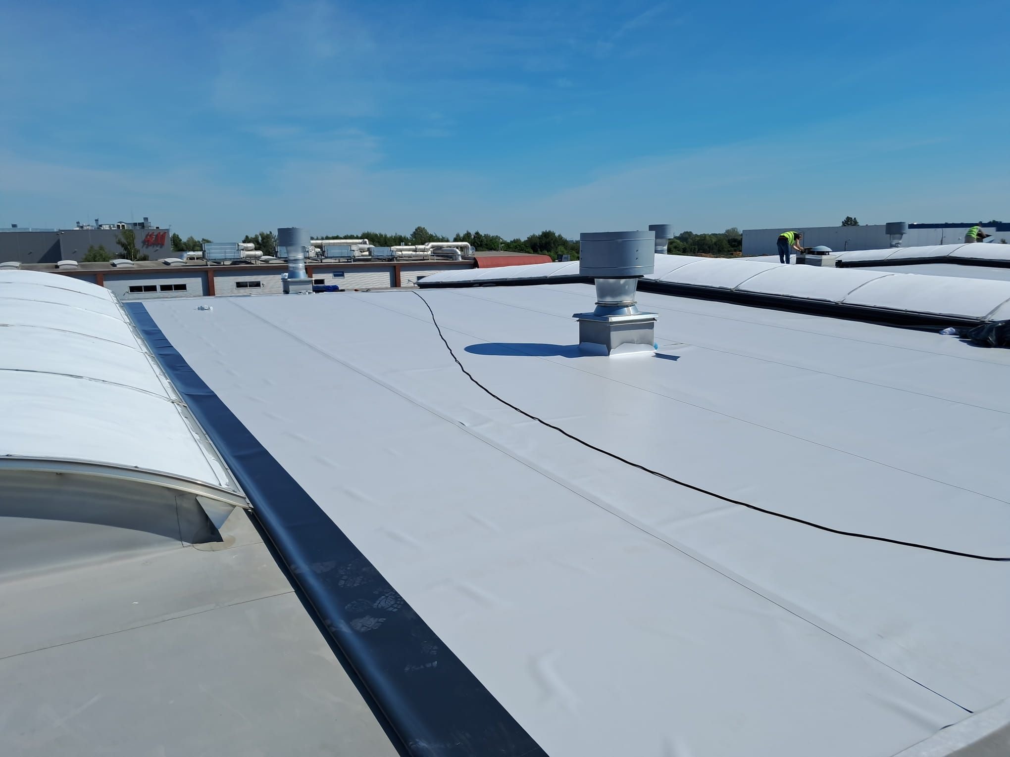 Jasnoszary, płaski dach przemysłowy z wentylatorami i czarnymi kablami, widoczne budynki w tle i fragment niebieskiego nieba.
