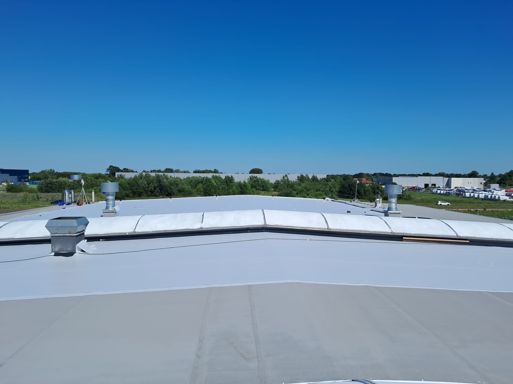 Szary dach przemysłowy z wentylacją, widok z góry, w tle zieleń i błękitne niebo.