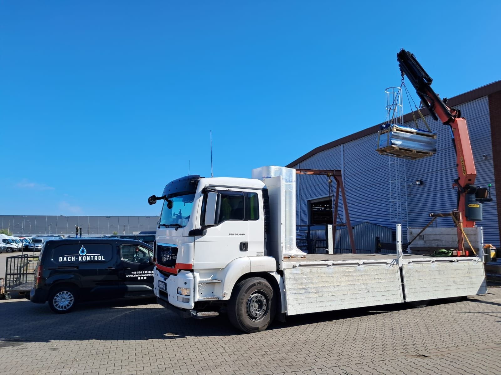 Ciężarówka z dźwigiem podnosi pakiet blachy trapezowej na tle magazynu z blachy falistej i firmowego busa z logo Dach-Kontrol.