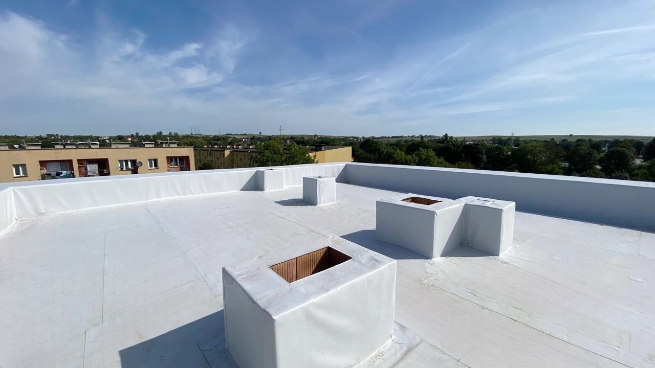 Biała, płaska powierzchnia dachu po hydroizolacji z kominami wentylacyjnymi, widok na zabudowania miejskie i błękitne niebo.