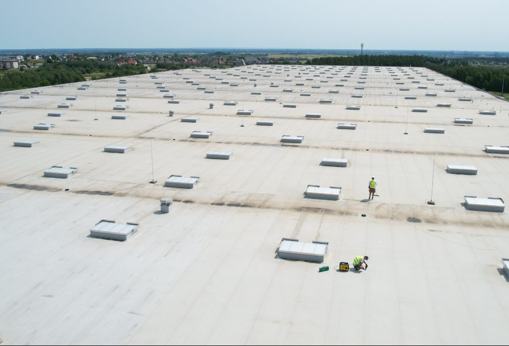 Rozległy, płaski dach przemysłowy z licznymi świetlikami; dwóch pracowników w odblaskowych kamizelkach wykonuje prace naprawcze na jasnej powierzchni dachu, widoczne narzędzia i agregat prądotwórczy.