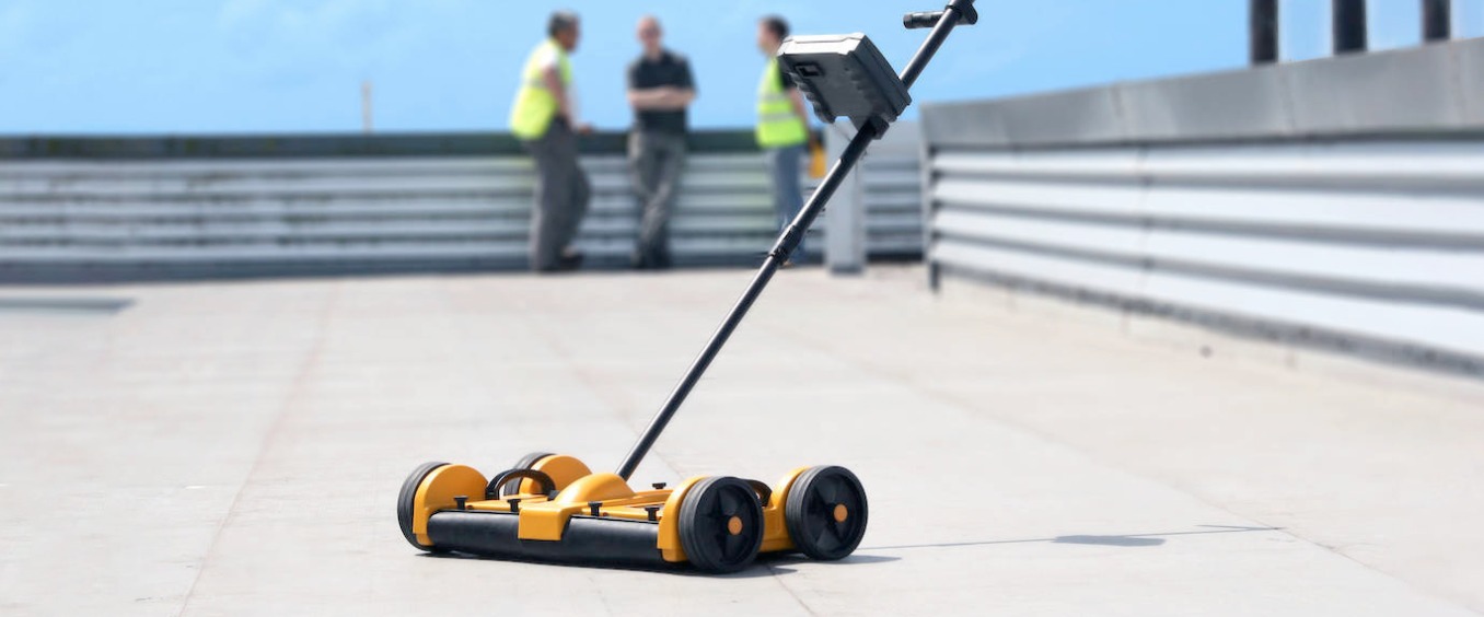 Żółto-czarne urządzenie diagnostyczne na dachu płaskim, z trzema osobami w odblaskowych kamizelkach w tle.