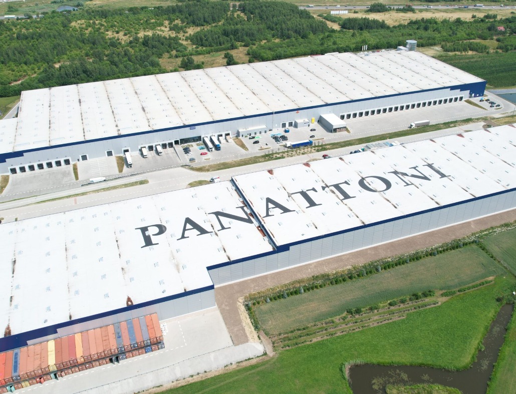 Widok z lotu ptaka na rozległy dach hali magazynowej z napisem 'PANATTONI', otoczony zielenią i innymi budynkami przemysłowymi.