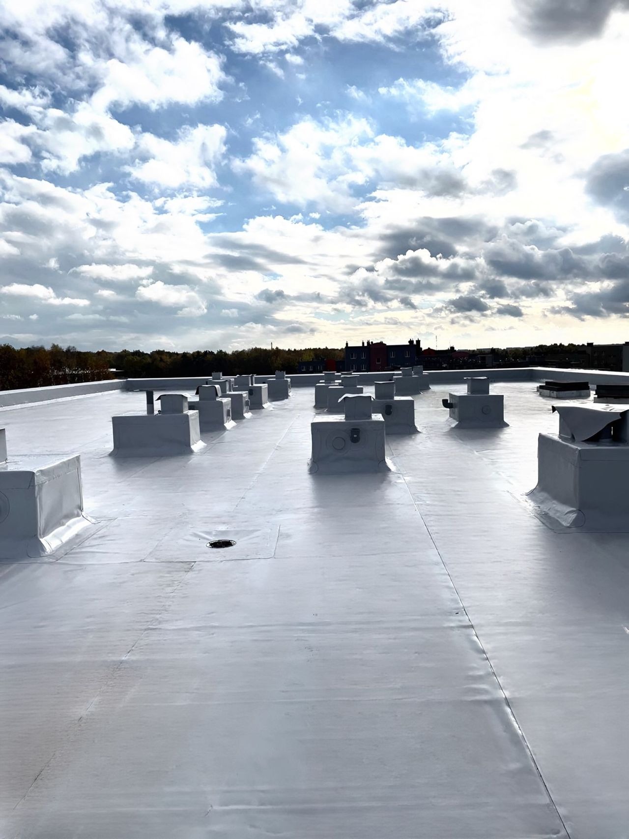Biała, płaska powierzchnia dachu z licznymi, równomiernie rozmieszczonymi odpowietrznikami, widoczne szwy łączenia membrany, na horyzoncie zieleń drzew i zabudowania miejskie, pochmurne niebo.