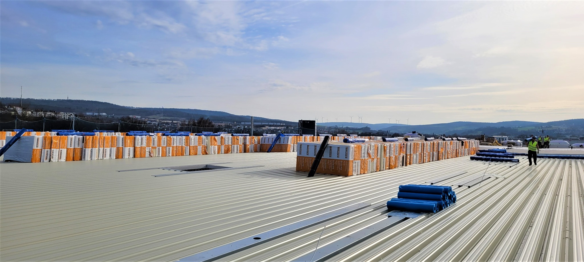 Rozległy, płaski dach przemysłowy w trakcie izolacji, z ułożonymi w rzędach pakietami materiałów izolacyjnych marki Bauder, zwiniętymi rolkami papy oraz pracownikami w odblaskowych kamizelkach,...