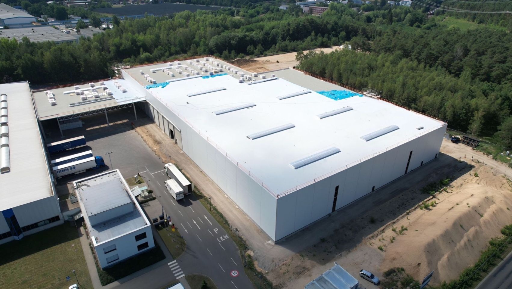 Widok z lotu ptaka na duży, biały dach hali przemysłowej z świetlikami, część dachu w trakcie prac wykończeniowych, obok zaparkowane ciężarówki.